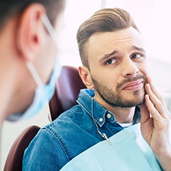 A young man sitting in a dentist’s chair holding his cheek before emergency dentistry